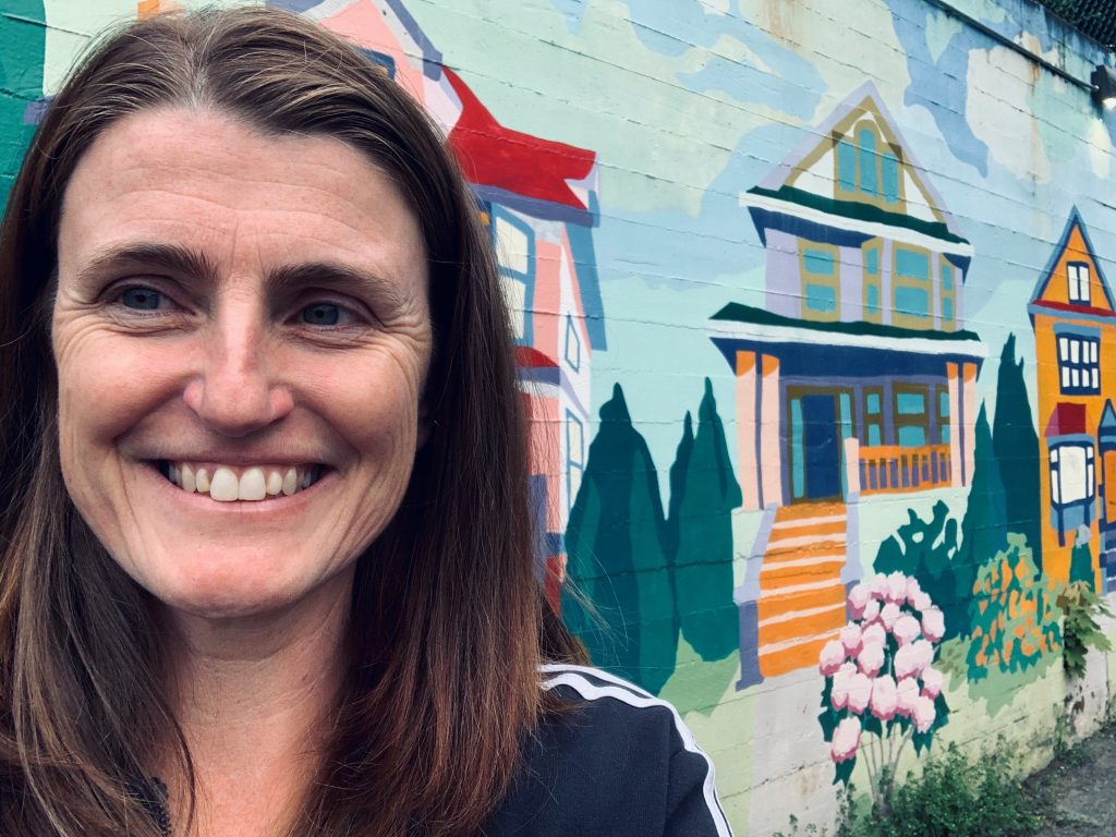 A happy adult white woman with a warm smile standing confidently in front of a vibrant and colourful wall mural.
